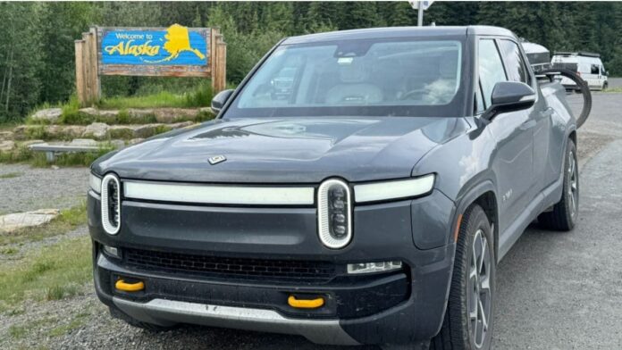 Gray Rivian R1T electric pickup truck parked by Alaska welcome sign, surrounded by forest landscape